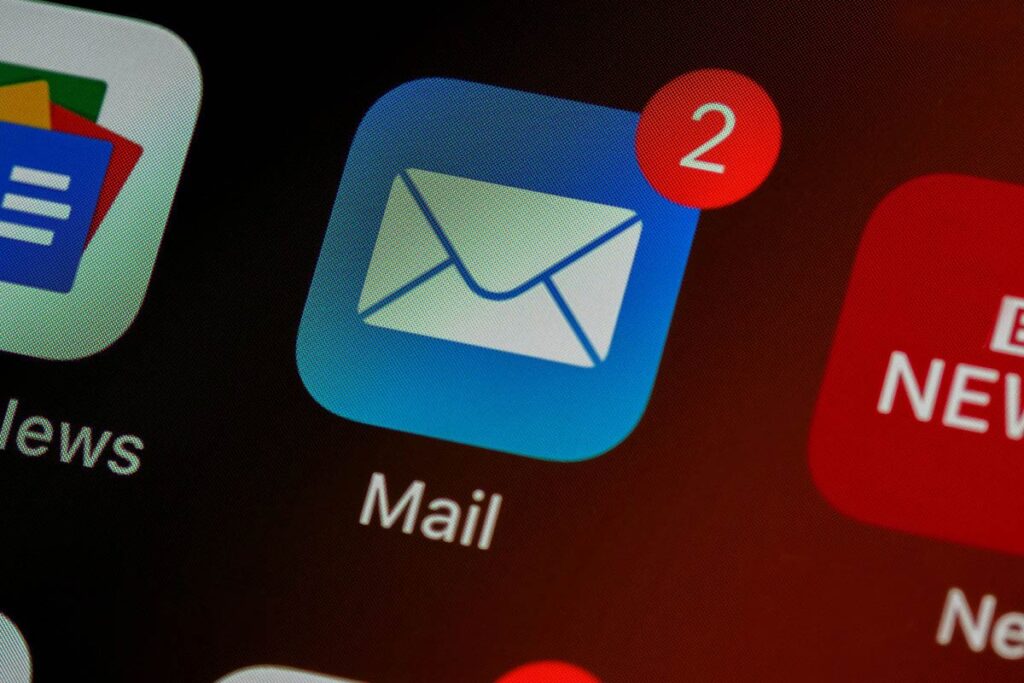 Tela de smartphone em close-up exibindo o ícone azul do aplicativo nativo de e-mail com a inscrição 'Mail' abaixo, destacando uma notificação de balão vermelho com o número dois no canto superior direito.