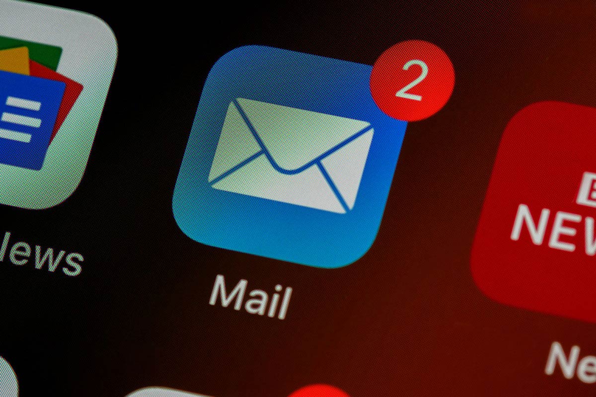 Tela de smartphone em close-up exibindo o ícone azul do aplicativo nativo de e-mail com a inscrição 'Mail' abaixo, destacando uma notificação de balão vermelho com o número dois no canto superior direito.