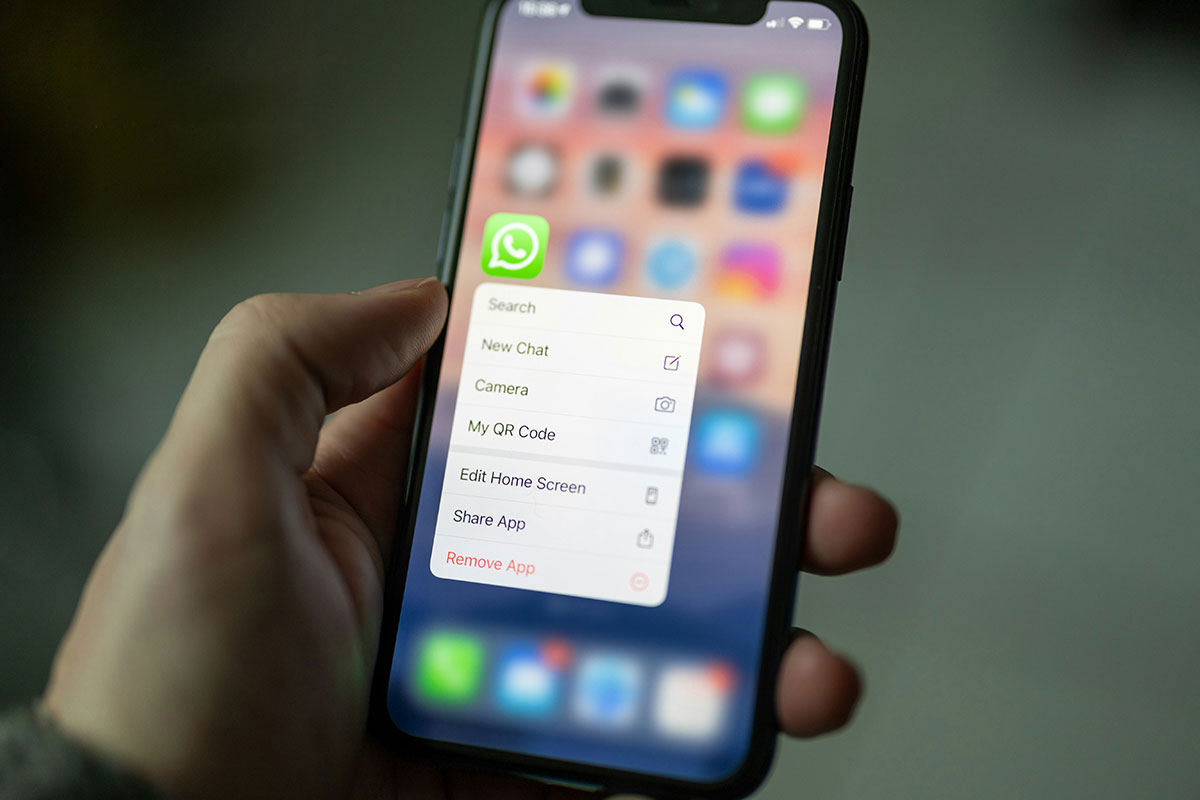 Mão humana segurando um smartphone em close-up, destacando o menu contextual pop-up aberto sobre o ícone verde do WhatsApp, exibindo opções de atalho como Nova Conversa e Meu Código QR.