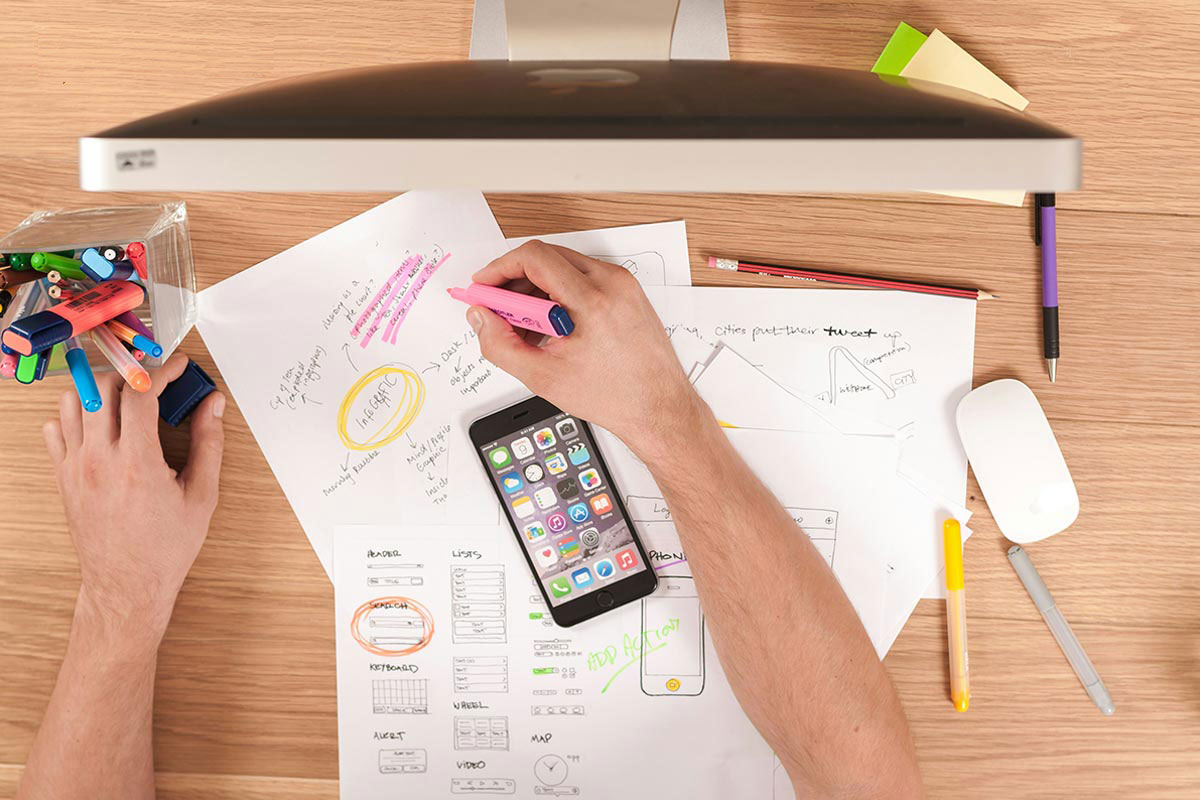 Visão aérea de um ambiente de trabalho em mesa de madeira, mostrando mãos que desenham e grifam wireframes, mapas mentais e infográficos em papéis sulfite ao lado de um smartphone e um monitor de computador.