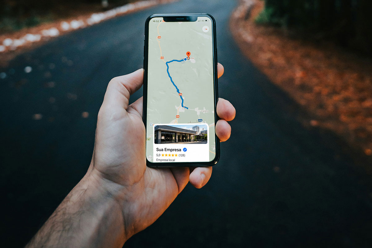 Mão humana segurando um smartphone em primeiro plano exibindo uma rota de navegação de mapa, com um card flutuante em destaque na tela mostrando a inscrição 'Sua Empresa', selo azul de verificação, avaliação cinco estrelas e a identificação 'Empresa local', tendo ao fundo uma estrada asfáltica desfocada.
