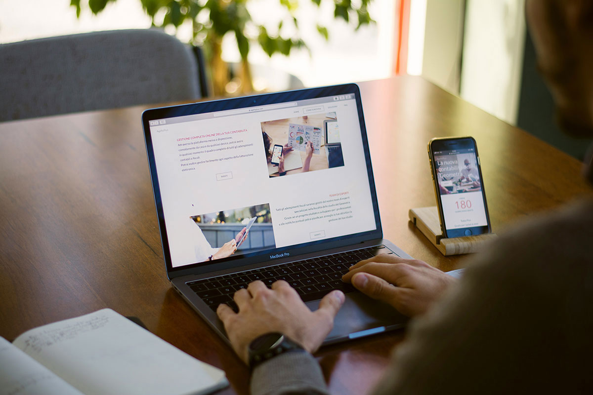Visão em perspectiva sobre o ombro de um homem usando um notebook MacBook sobre uma mesa de madeira, com um smartphone ao lado em um suporte de madeira exibindo uma página mobile.