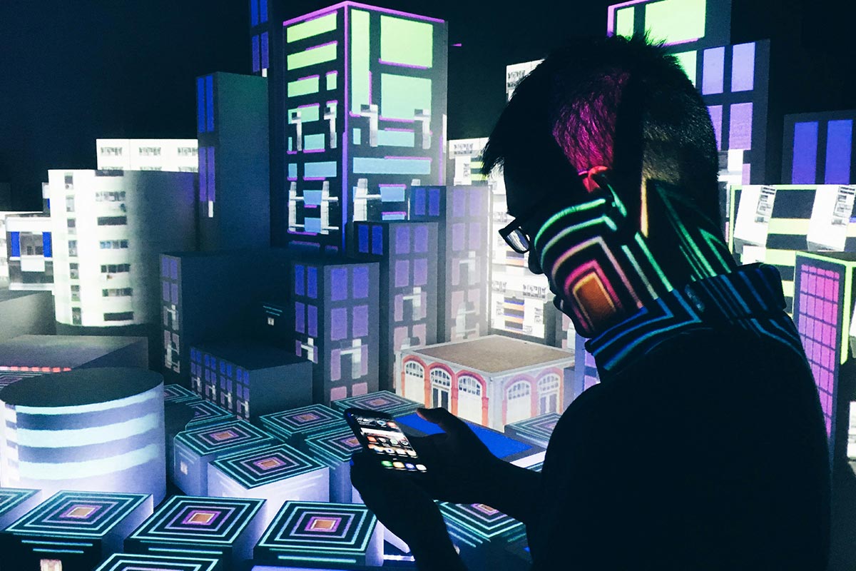 Homem de perfil olha para a tela de seu smartphone em um ambiente escuro e futurista iluminado por projeções coloridas de prédios e cubos geométricos digitais.