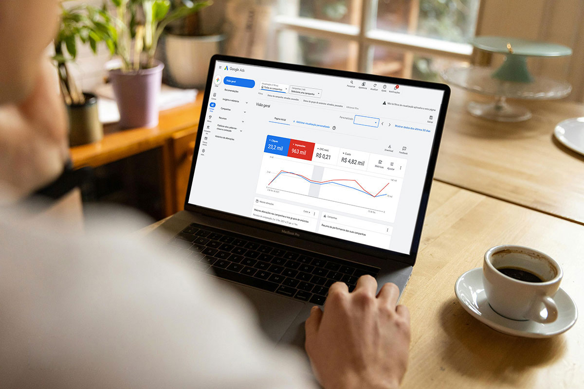 Visão em primeira pessoa de uma pessoa sentada a uma mesa de madeira, tomando café enquanto utiliza um notebook MacBook Pro que exibe o painel de visão geral do Google Ads, mostrando métricas de cliques, impressões, CPC médio e custo total.