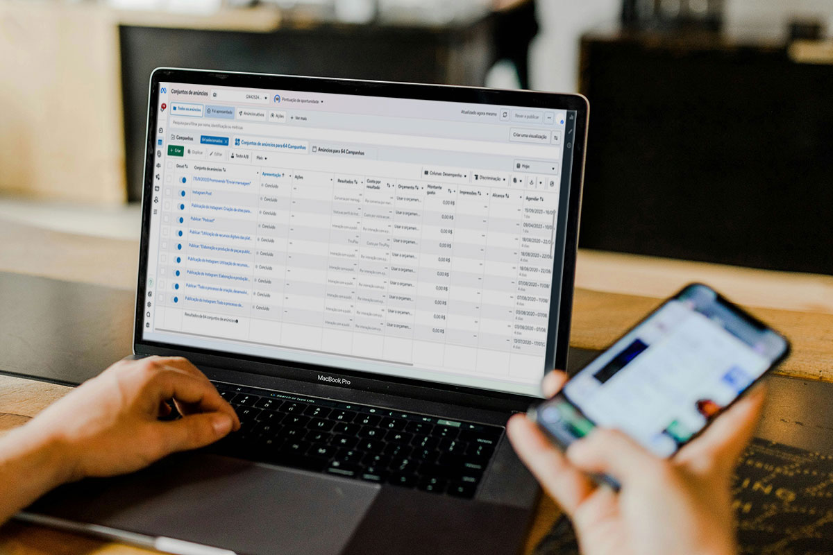 Visão em primeira pessoa de uma pessoa trabalhando em uma mesa de madeira, com as mãos sobre um MacBook Pro exibindo o painel detalhado do Gerenciador de Anúncios da Meta, enquanto segura um smartphone na outra mão.