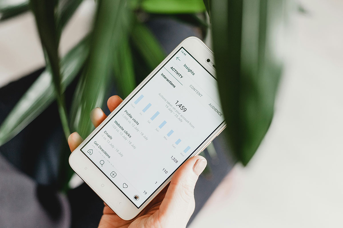 Mão segurando um smartphone branco que exibe uma tela de métricas e gráficos de dados analíticos intitulada 'Insights', com um fundo desfocado de folhas verdes.