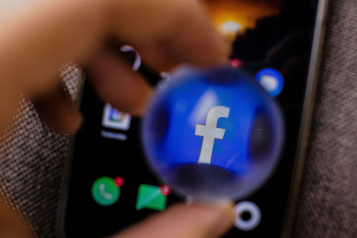 Uma mão segura uma lente de aumento esférica diante da tela de um smartphone, ampliando o ícone azul e branco do aplicativo do Facebook em destaque, com o fundo da tela levemente desfocado.