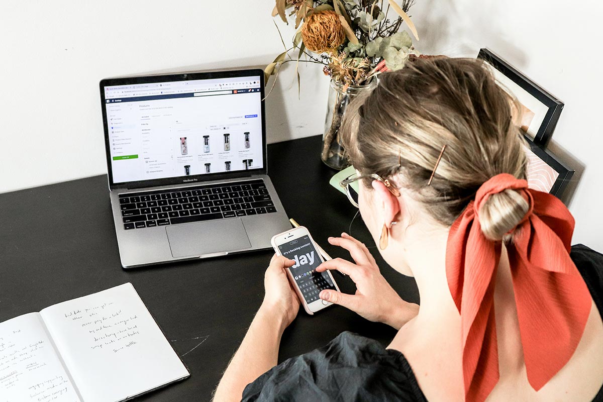 Visão aérea de uma mulher trabalhando em uma mesa preta, utilizando um notebook que exibe uma lista de produtos cadastrados para e-commerce e, simultaneamente, segurando um smartphone com as mãos para gerenciar conteúdos digitais.