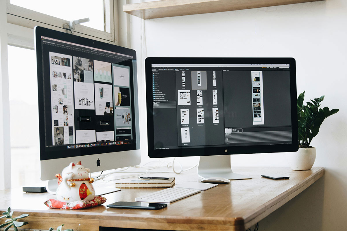 Dois monitores de computador de alta resolução posicionados sobre uma mesa de madeira clara, exibindo layouts de design de websites em processo de criação, com um pequeno boneco da sorte japonês e uma planta ao lado.
