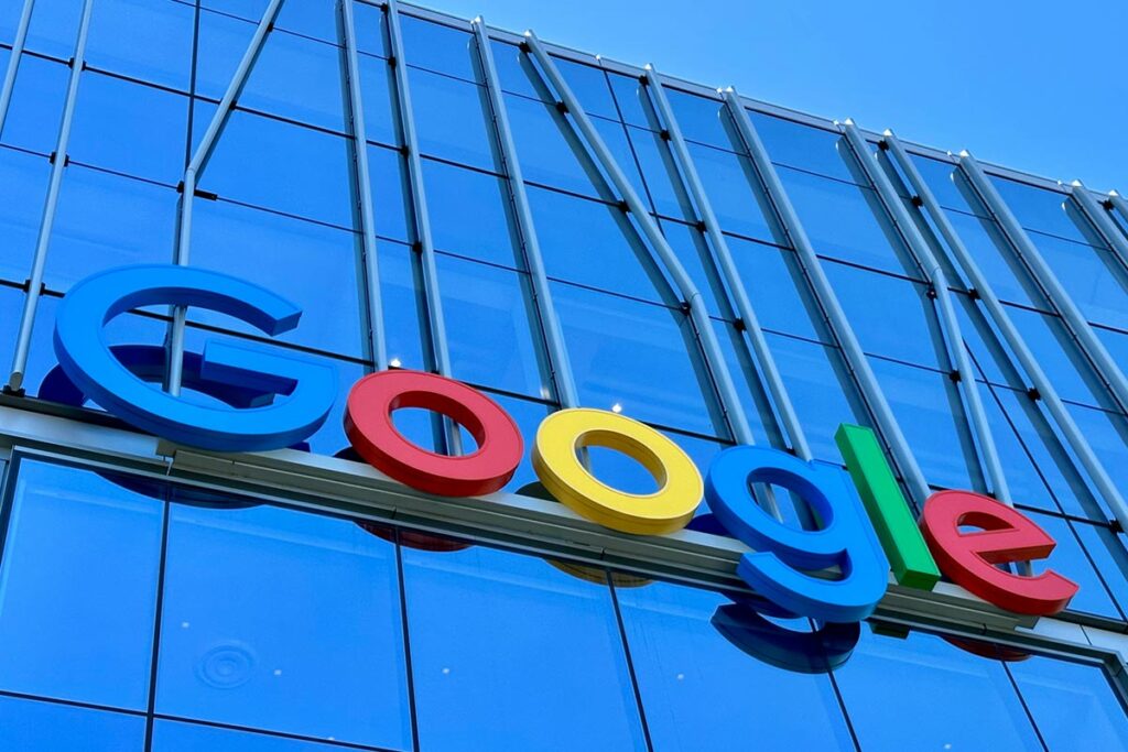 Logotipo tridimensional colorido do Google (letras G azul, o vermelho, o amarelo, g azul, l verde e e vermelho) fixado na fachada moderna e espelhada de um edifício de vidro azul sob um céu limpo.