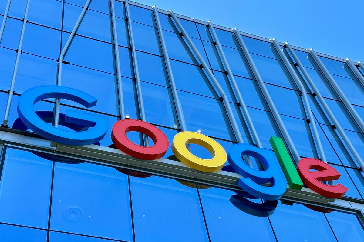 Logotipo tridimensional colorido do Google (letras G azul, o vermelho, o amarelo, g azul, l verde e e vermelho) fixado na fachada moderna e espelhada de um edifício de vidro azul sob um céu limpo.