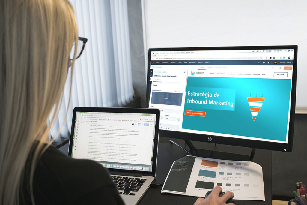 Mulher loira vista de costas trabalhando em uma mesa de escritório com um notebook aberto e olhando para um monitor de desktop maior que exibe um construtor de páginas com o título Estratégia de Inbound Marketing ao lado do desenho de um funil de vendas colorido.