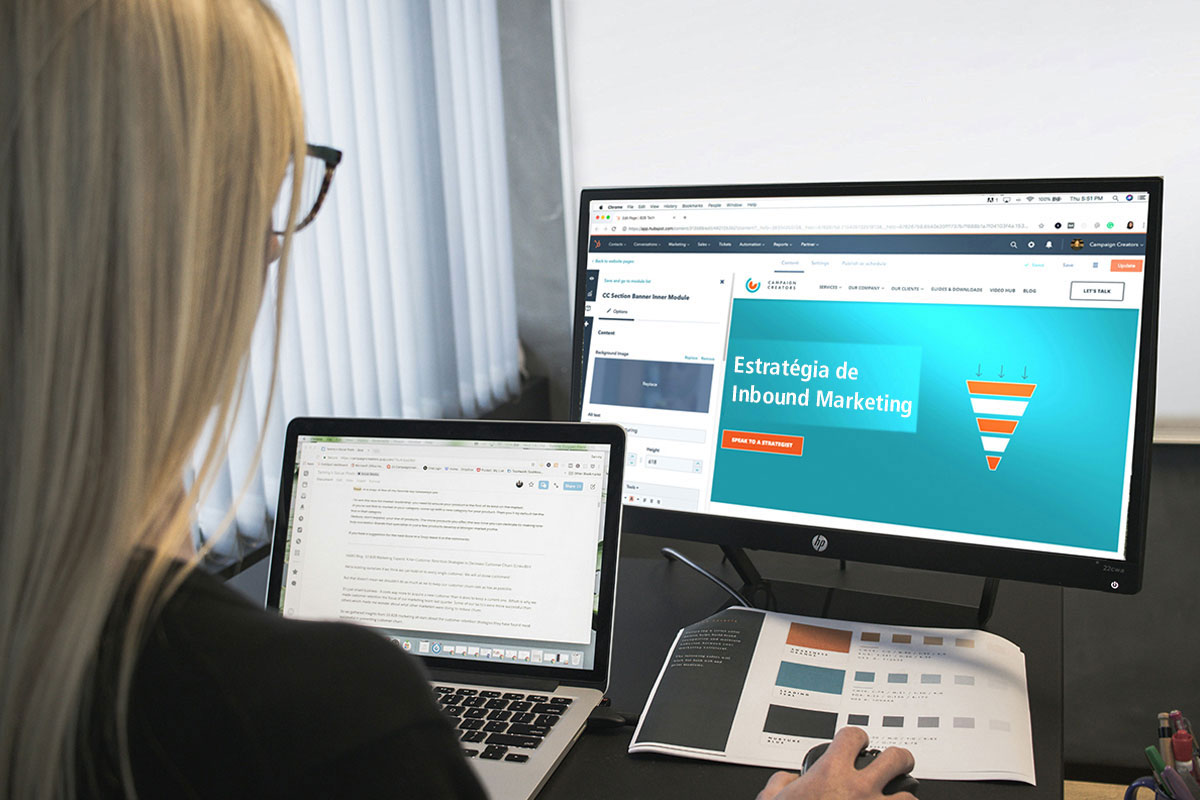 Mulher loira vista de costas trabalhando em uma mesa de escritório com um notebook aberto e olhando para um monitor de desktop maior que exibe um construtor de páginas com o título Estratégia de Inbound Marketing ao lado do desenho de um funil de vendas colorido.