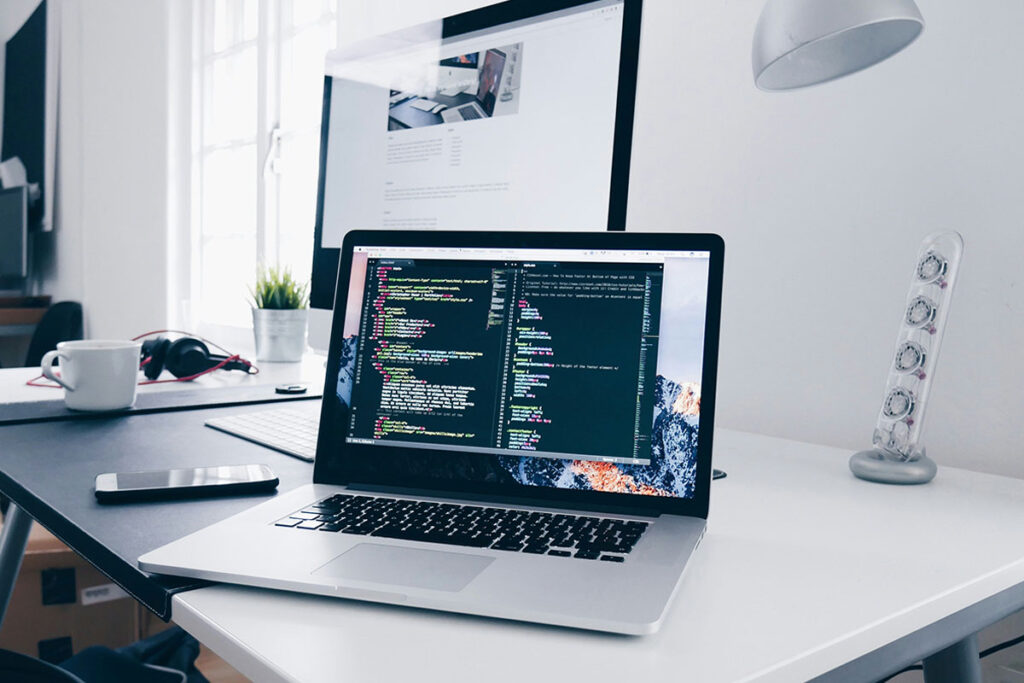 Notebook em primeiro plano exibindo linhas de código de programação em uma mesa de escritório branca e organizada, com monitor de desktop ao fundo, representando a infraestrutura técnica da API de Conversões.