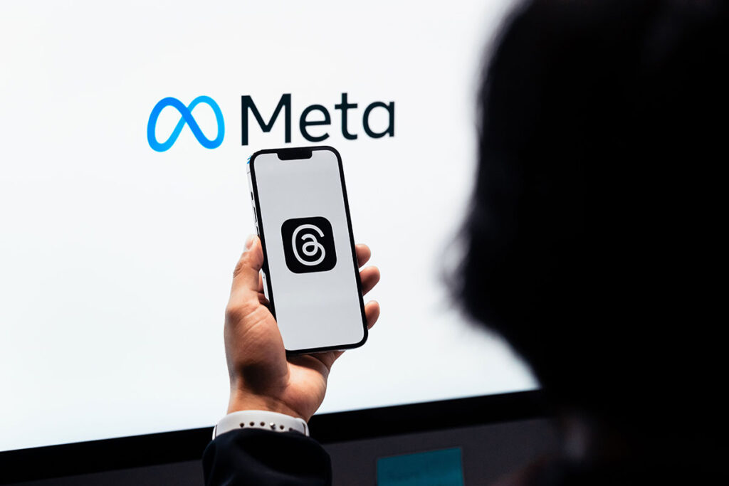 Uma pessoa segura um smartphone em primeiro plano mostrando um aplicativo em uma tela clara, com o logotipo azul e a palavra Meta exibidos ao fundo em uma tela branca.