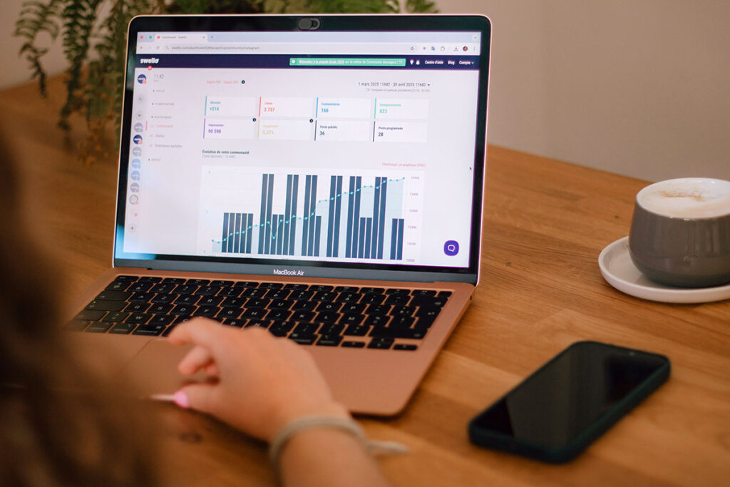 Visão em plano fechado sobre o ombro de uma pessoa analisando um painel de métricas de marketing digital com gráficos de barras verticais e linhas de conversão na tela de um notebook MacBook Air sobre uma mesa de madeira clara.