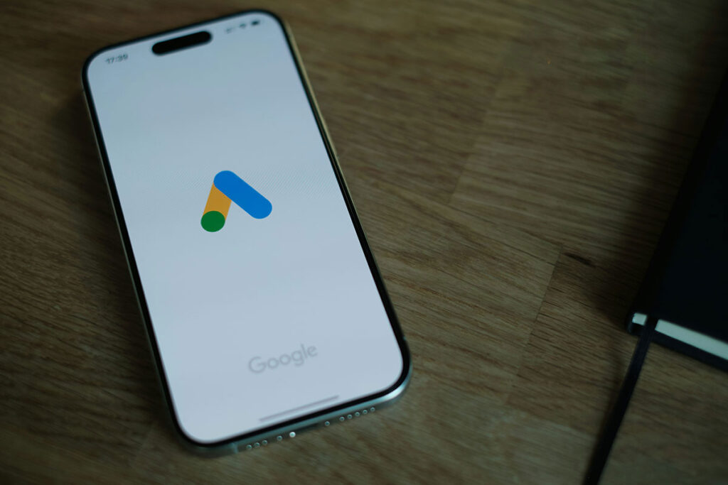 Smartphone com a tela ligada repousando sobre uma mesa de madeira, exibindo o logotipo colorido do Google Ads centralizado.