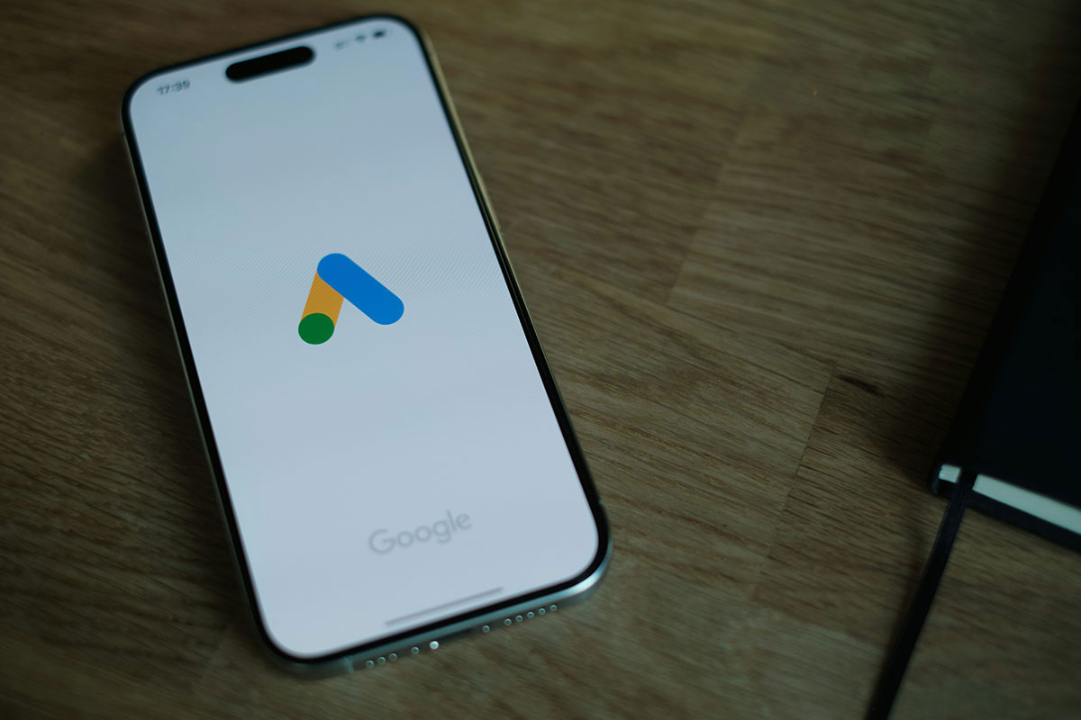 Smartphone com a tela ligada repousando sobre uma mesa de madeira, exibindo o logotipo colorido do Google Ads centralizado.