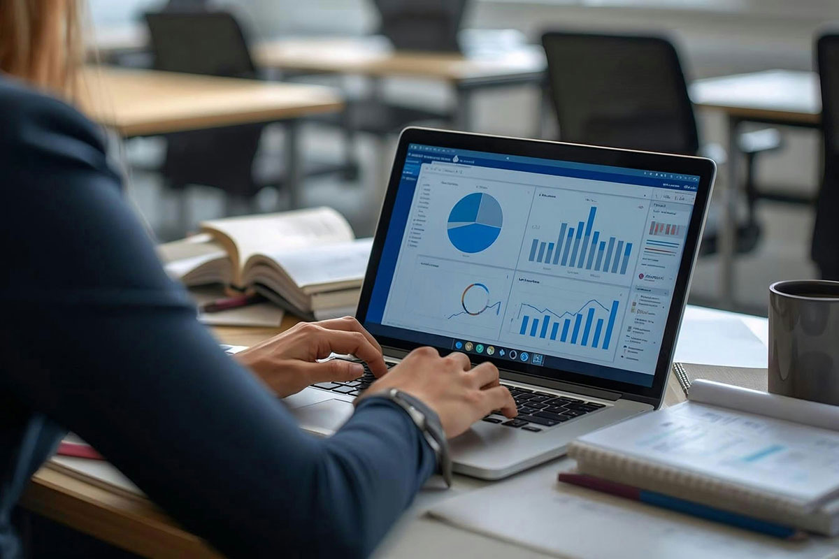 Uma pessoa trabalhando em um notebook que exibe diversos gráficos de análise de dados e painéis de performance financeira, sentada em uma mesa de escritório organizada com cadernos.