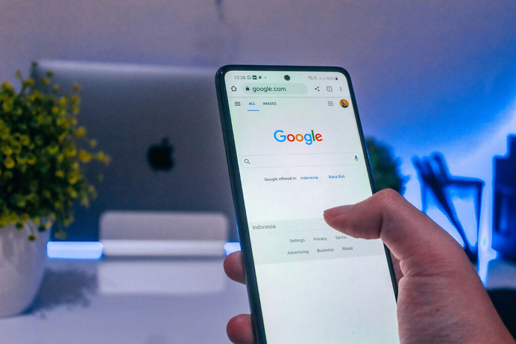 Mão segurando um smartphone que exibe a página inicial do buscador Google em um navegador mobile, com um notebook desfocado ao fundo em um ambiente com iluminação LED azul.