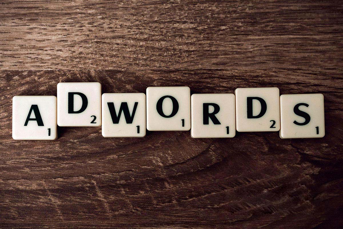 Peças de jogo de tabuleiro estilo Scrabble formando a palavra 'ADWORDS' sobre uma superfície de madeira escura.