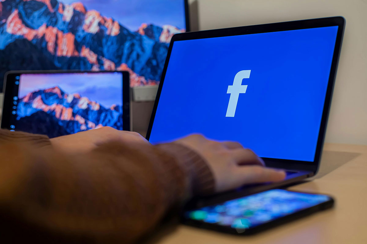 Visão lateral de uma pessoa trabalhando em um notebook que exibe o logotipo do Facebook na tela, com um smartphone desfocado em primeiro plano sobre a mesa de madeira.