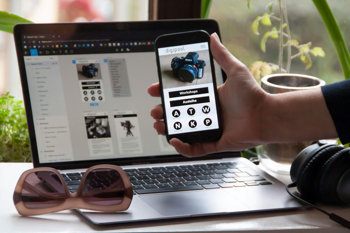 Mão segurando um smartphone que exibe a interface de um site, posicionada à frente de um notebook que mostra o software de design de layout de páginas (Figma) em uma mesa de trabalho organizada, demonstrando o conceito de design responsivo focado no celular.