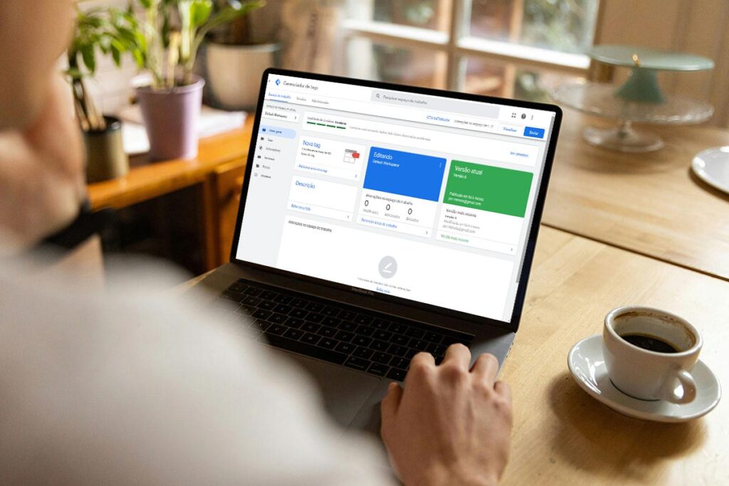 Foto em ângulo oblíquo traseiro de uma pessoa usando um MacBook Pro em uma mesa de madeira clara. A tela do notebook exibe a página inicial do Google Tag Manager. Há uma xícara de café à direita e plantas ao fundo em um ambiente bem iluminado.
