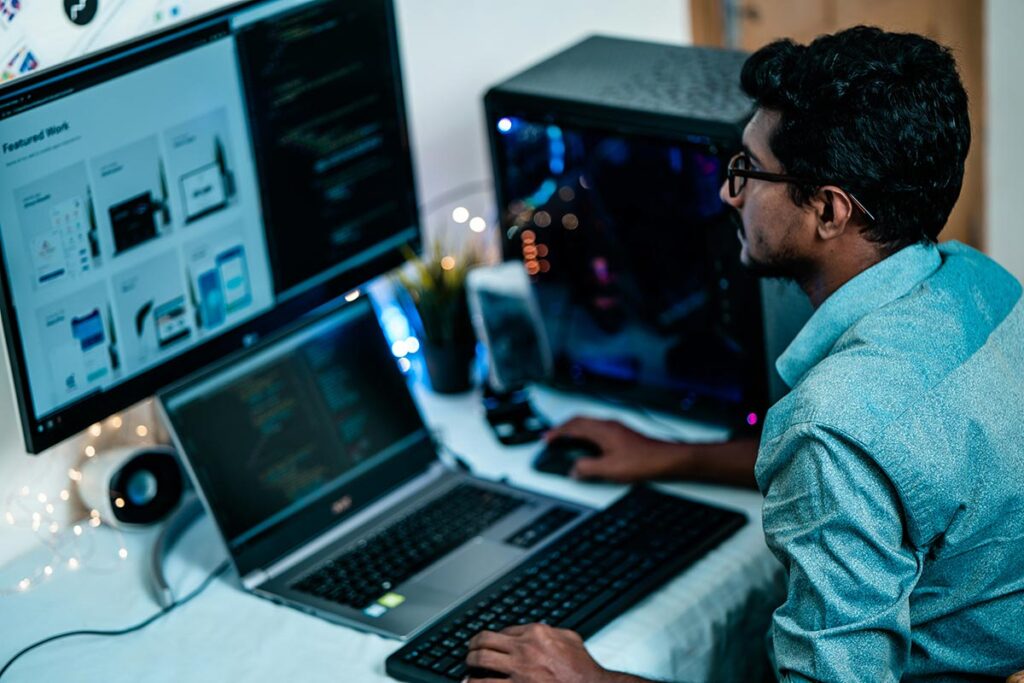 Desenvolvedor de perfil trabalhando em sua mesa com um monitor grande exibindo layouts de páginas web em mosaico e linhas de código, ao lado de um notebook aberto com telas de programação.