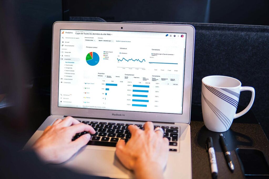 Visão em primeira pessoa de mãos digitando em um notebook MacBook que exibe um painel com gráficos de pizza e tabelas de métricas de tráfego web, com uma caneca de café ao lado sobre a mesa.