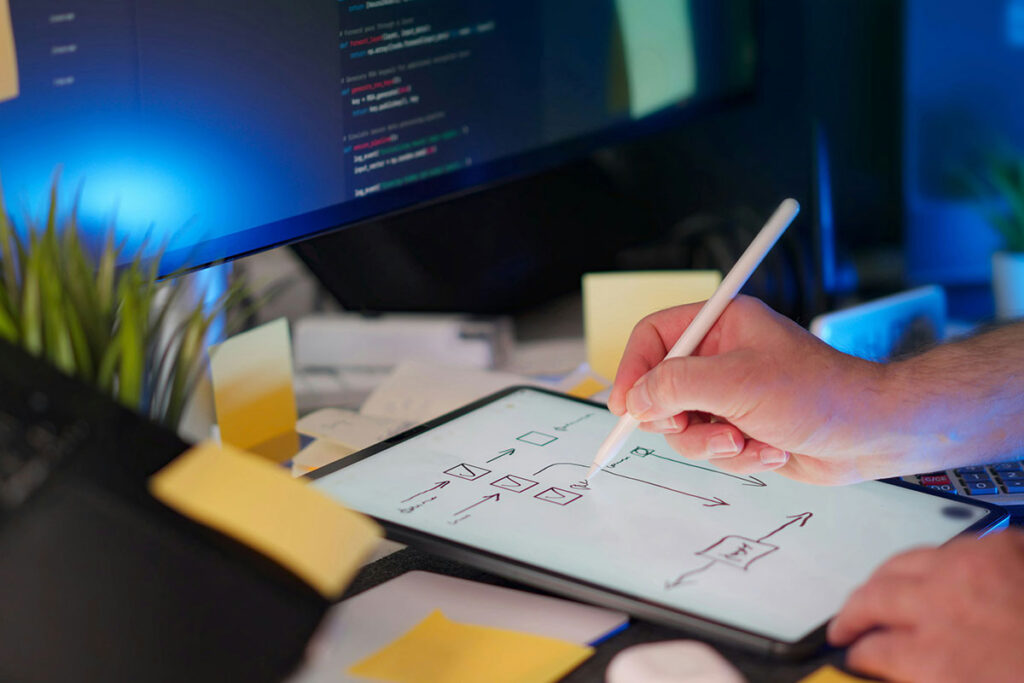 Mão segurando uma caneta digital e desenhando um fluxograma estratégico com setas e blocos na tela de um tablet, sobre uma mesa de trabalho com blocos de notas amarelos e monitor ao fundo exibindo linhas de código sob luz azulada.