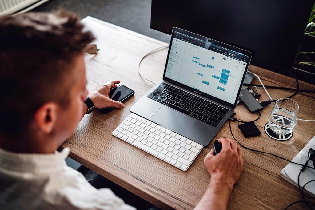 Visão aérea de um analista de marketing trabalhando em uma mesa de madeira. Ele usa a mão esquerda para segurar um smartphone e a mão direita em um mouse sem fio ao lado de um teclado externo. Na tela do notebook, é exibida uma interface de calendário e cronograma de projetos.