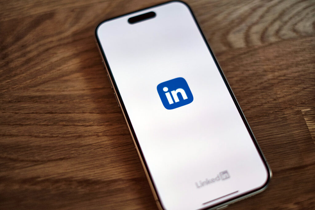 Tela de um smartphone iPhone sobre uma mesa de madeira rústica, exibindo o ícone azul da rede social LinkedIn em fundo branco de forma minimalista.