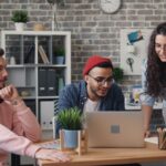 Equipe de profissionais de marketing em um escritório com parede de tijolos reunida ao redor de uma mesa de madeira, analisando o planejamento de anúncios na tela de um notebook.