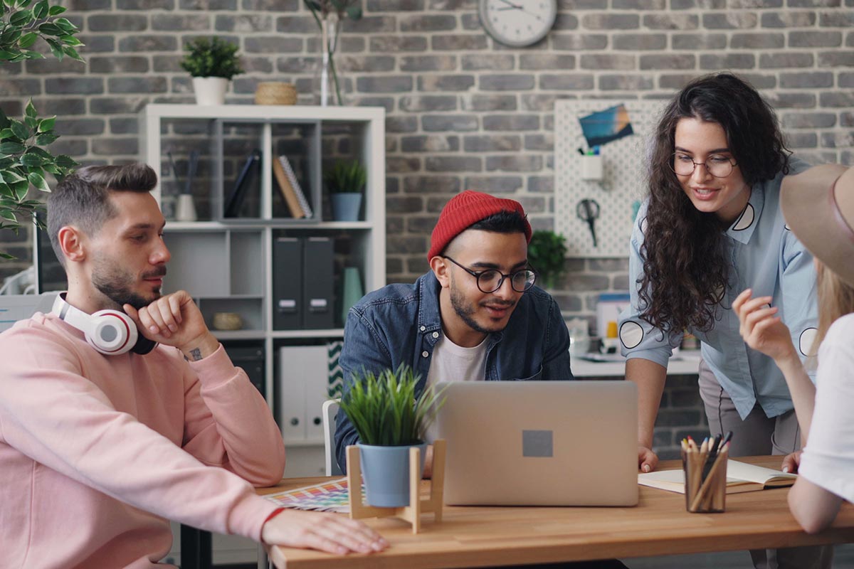 Equipe de profissionais de marketing em um escritório com parede de tijolos reunida ao redor de uma mesa de madeira, analisando o planejamento de anúncios na tela de um notebook.