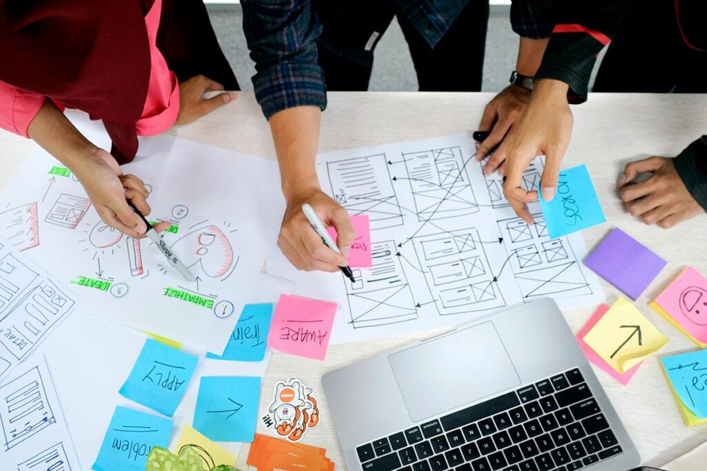 Equipe de UX design planejando a arquitetura de informação de um site com wireframes e post-its em uma mesa de trabalho.