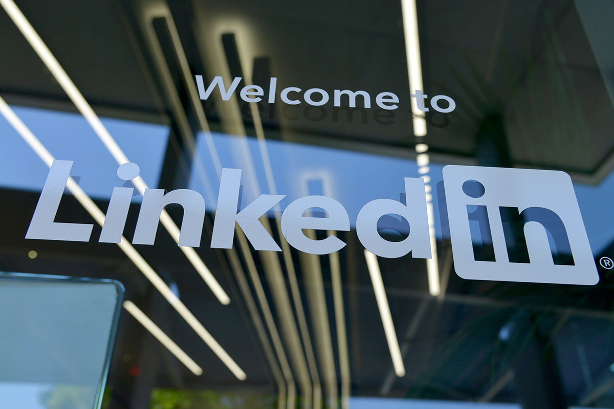Logotipo do LinkedIn em letras brancas colado na vidraça da recepção de um escritório moderno corporativo, com a frase "Welcome to" acima do nome da plataforma.