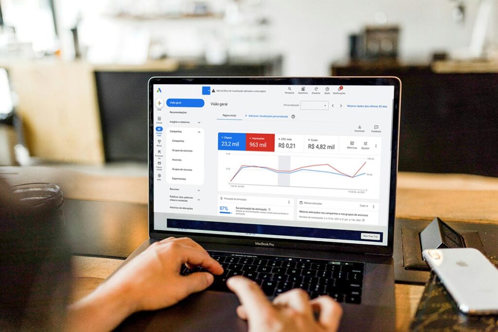 Visão em primeiro plano das mãos de um profissional digitando no teclado de um notebook. A tela mostra o painel de visão geral do Google Ads em português, exibindo gráficos de desempenho com métricas de cliques, impressões, CPC médio e custo total de campanhas de tráfego pago.