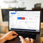 Visão em primeiro plano das mãos de um profissional digitando no teclado de um notebook. A tela mostra o painel de visão geral do Google Ads em português, exibindo gráficos de desempenho com métricas de cliques, impressões, CPC médio e custo total de campanhas de tráfego pago.
