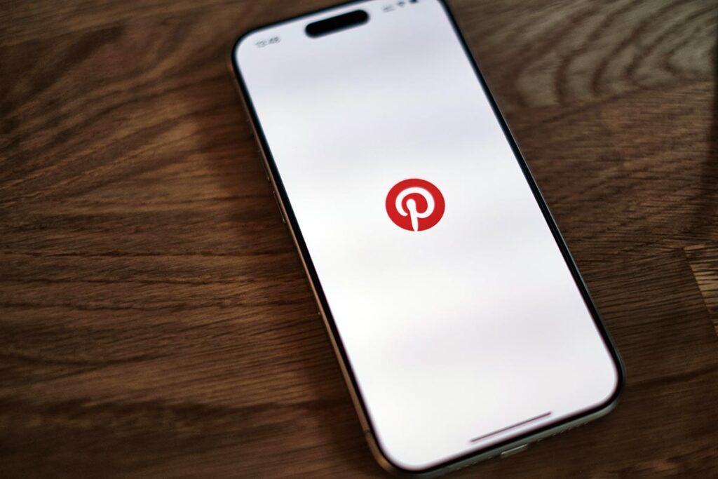 Smartphone sobre mesa de madeira com o aplicativo do Pinterest aberto exibindo o logotipo vermelho da plataforma centralizado na tela.
