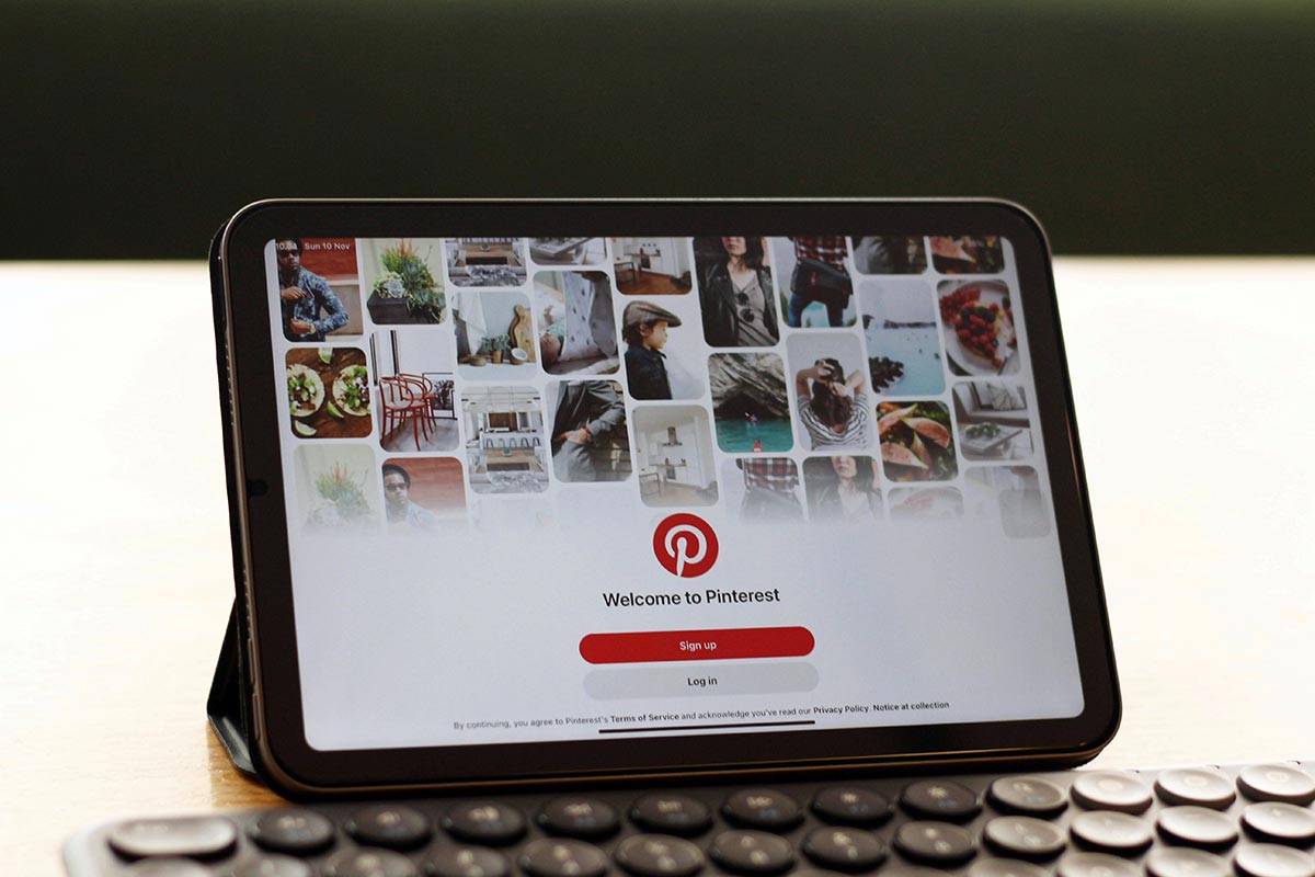 Tablet sobre uma mesa de madeira exibindo a tela inicial do aplicativo Pinterest com um mosaico de Pins visuais e o logotipo centralizado.