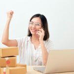 Mulher asiática de óculos, sorrindo e comemorando com o punho levantado, sentada à mesa de madeira com um notebook prateado e caixas de papelão empilhadas.