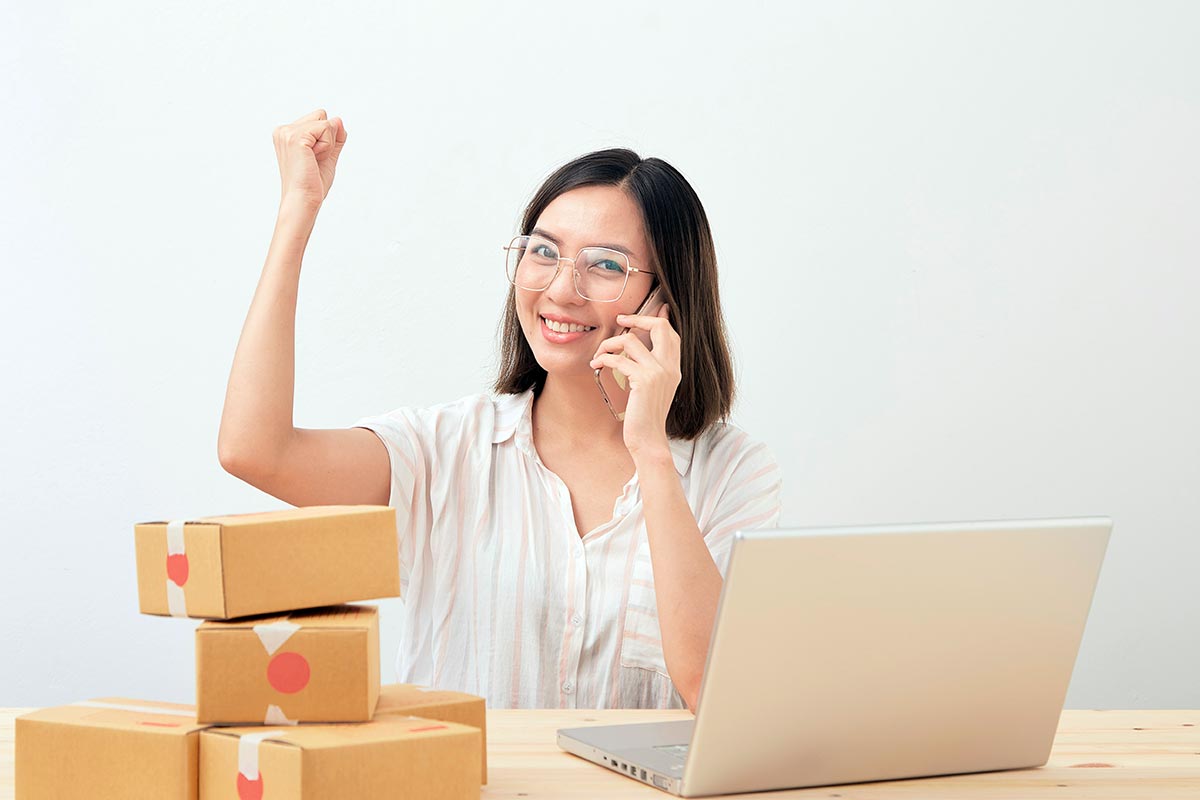 Mulher asiática de óculos, sorrindo e comemorando com o punho levantado, sentada à mesa de madeira com um notebook prateado e caixas de papelão empilhadas.