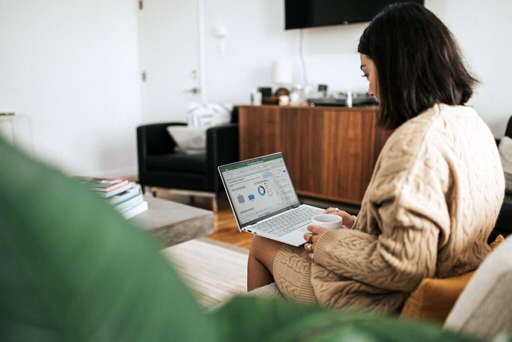 Visão por trás e lateral de uma mulher sentada em um sofá, segurando uma xícara, enquanto trabalha em um notebook prateado que exibe gráficos de barras e pizzas em uma planilha de acompanhamento mensal.