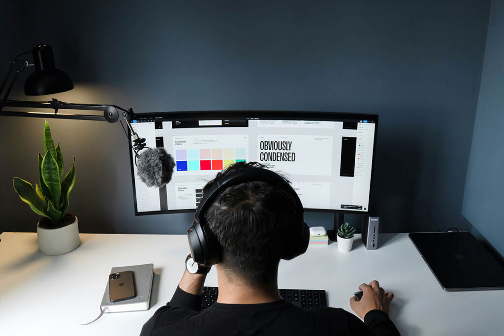 Visão em perspectiva traseira de um designer com fones de ouvido analisando uma interface de site em um monitor ultra-wide, destacando uma paleta de cores quadradas e elementos de tipografia.