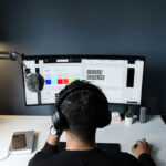Visão em perspectiva traseira de um designer com fones de ouvido analisando uma interface de site em um monitor ultra-wide, destacando uma paleta de cores quadradas e elementos de tipografia.