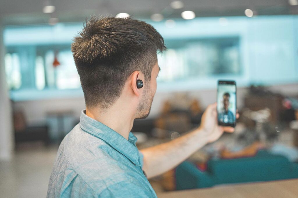 Homem de camisa azul clara, visto de costas e perfil, segurando um smartphone na vertical para gravar um vídeo, em um ambiente de escritório com iluminação natural.