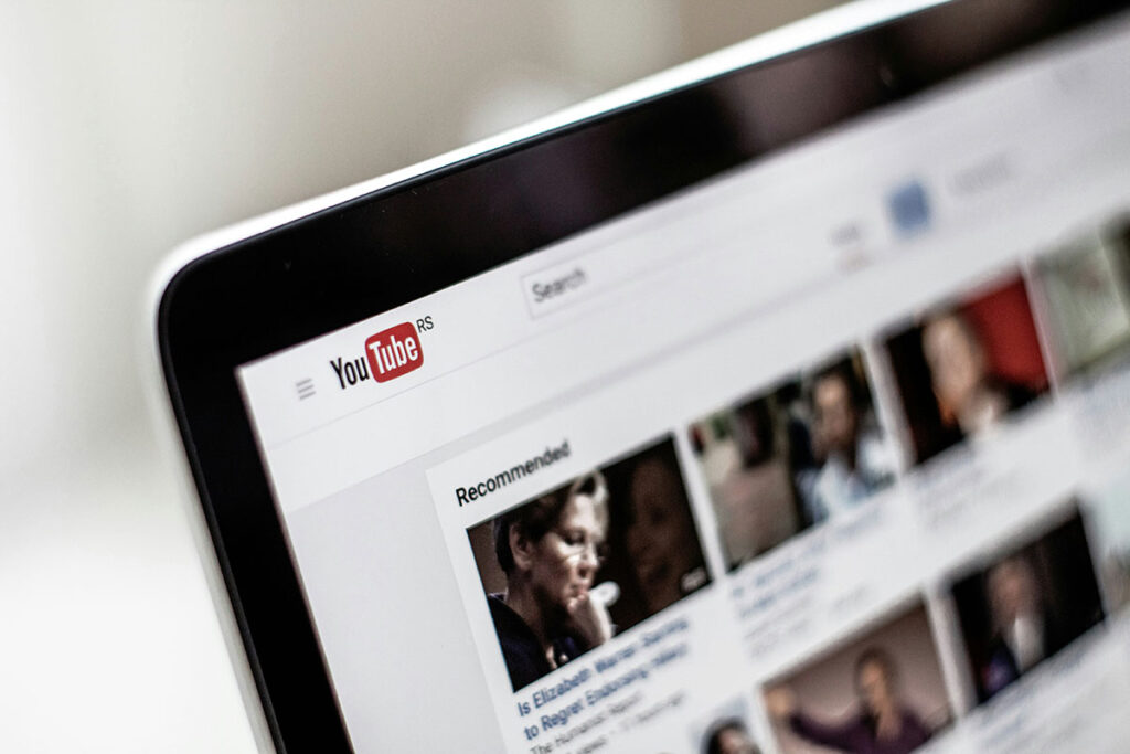 Notebook em ângulo exibindo a página inicial do YouTube com vídeos sugeridos na tela, com foco seletivo e fundo claro desfocado.