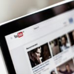 Notebook em ângulo exibindo a página inicial do YouTube com vídeos sugeridos na tela, com foco seletivo e fundo claro desfocado.