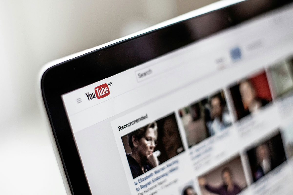 Notebook em ângulo exibindo a página inicial do YouTube com vídeos sugeridos na tela, com foco seletivo e fundo claro desfocado.